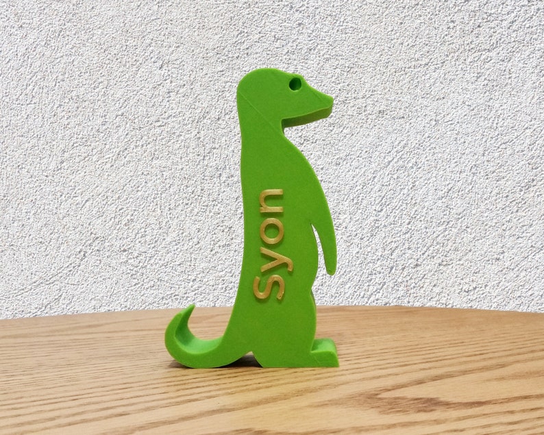 K&ouml;nnte beinhalten: Eine gr&uuml;ne Plastikfigur eines Erdm&auml;nnchens, das auf einer Holzoberfl&auml;che steht. Das Erdm&auml;nnchen schaut nach rechts und hat das Wort "S'yon" in goldenen Buchstaben auf der Seite geschrieben.
