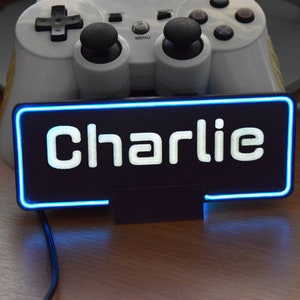 Pu&ograve; includere: Un cartello nero con un contorno al neon blu e la parola "Charlie" in lettere bianche. Il cartello &egrave; appoggiato su una superficie di legno.