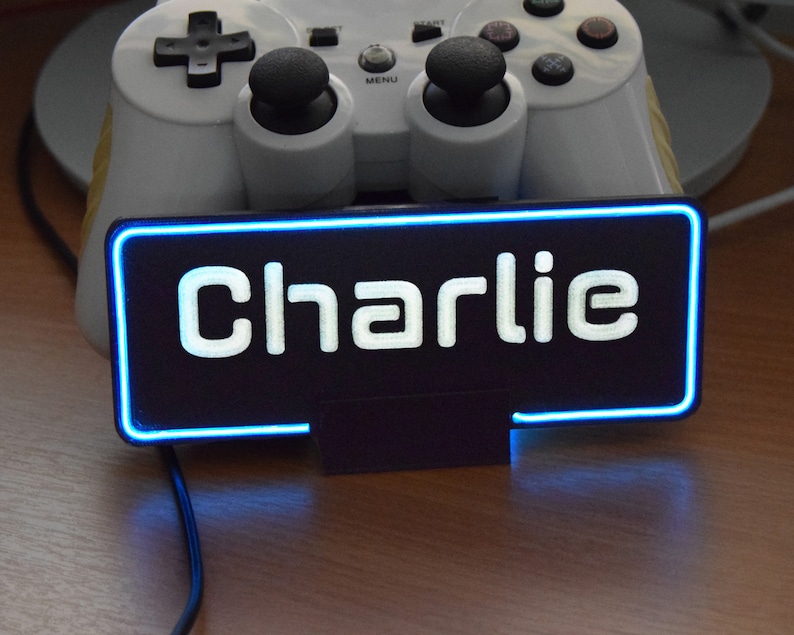 Pu&ograve; includere: Un cartello nero con un contorno al neon blu e la parola "Charlie" in lettere bianche. Il cartello &egrave; appoggiato su una superficie di legno.