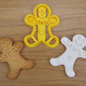 Puede incluir: Dos galletas de hombre de jengibre, una con el nombre "JAMES" y la otra con el nombre "EMMA", y un cortador de galletas de hombre de jengibre de pl&aacute;stico amarillo.