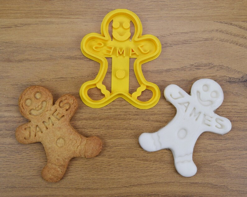 Puede incluir: Dos galletas de hombre de jengibre, una con el nombre "JAMES" y la otra con el nombre "EMMA", y un cortador de galletas de hombre de jengibre de pl&aacute;stico amarillo.