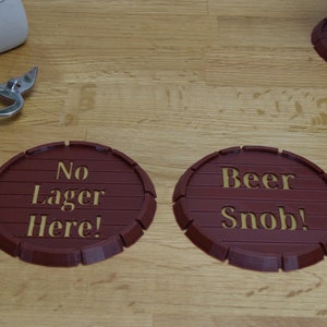 May include: Two brown, barrel-shaped coasters with gold lettering. One reads "No Lager Here!" and the other says "Beer Snob!". A bottle opener and a glass of beer are also visible.