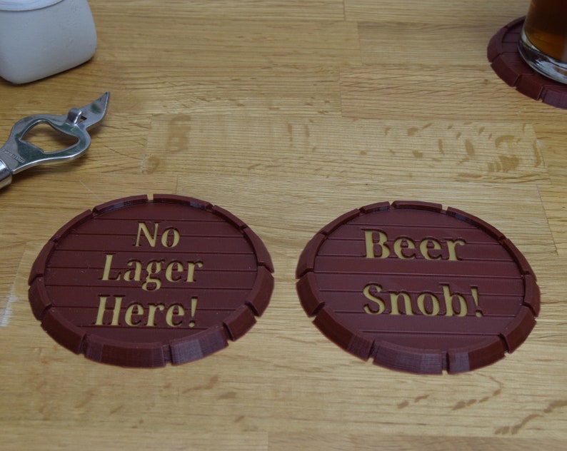 May include: Two brown, barrel-shaped coasters with gold lettering. One reads "No Lager Here!" and the other says "Beer Snob!". A bottle opener and a glass of beer are also visible.