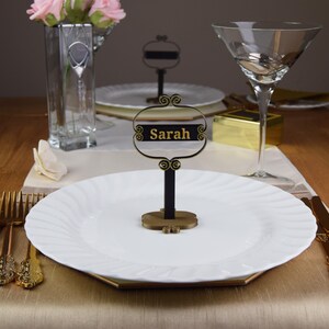 May include: A black and gold table place card with the name "Sarah" written in gold lettering. The place card is on a white plate with a gold rim.
