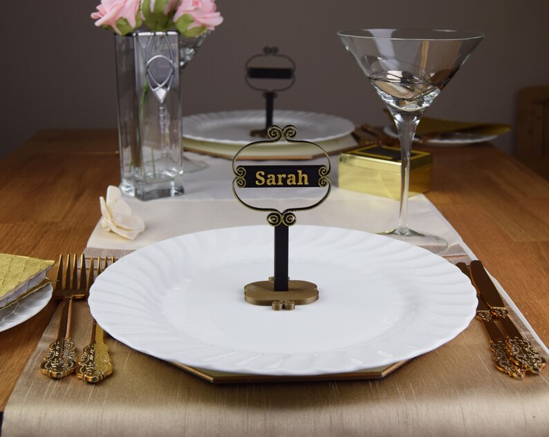 May include: A black and gold table place card with the name "Sarah" written in gold lettering. The place card is on a white plate with a gold rim.