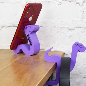 May include: Two purple 3D printed phone stands shaped like cartoon snakes. One snake is holding a red iPhone. The snake holding the phone has the name "Sophie" printed on it.