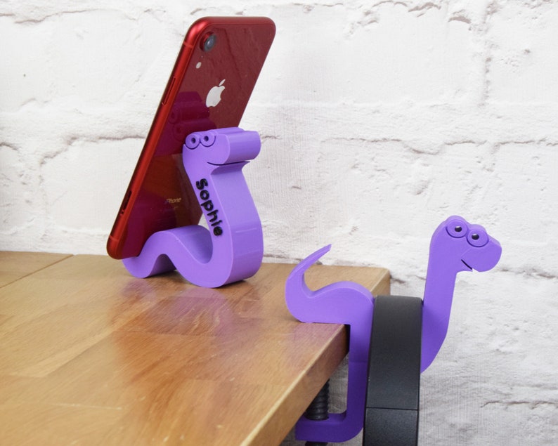May include: Two purple 3D printed phone stands shaped like cartoon snakes. One snake is holding a red iPhone. The snake holding the phone has the name "Sophie" printed on it.
