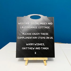 May include: Black chalkboard sign with white text that reads "Welcome to Guernsey and La Cordarge Cottage. Please enjoy these complimentary items on us. Warm wishes, Matthew and Tania X".
