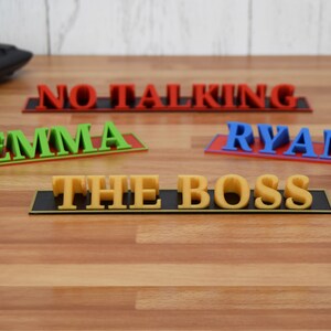 May include: A collection of colorful desk nameplates and signs. The signs include the names "Emma" in green, "Ryan" in blue, and the phrase "The Boss" in yellow. A red sign reads "No Talking".