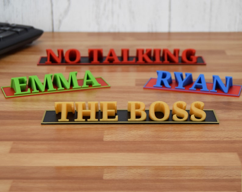 May include: A collection of colorful desk nameplates and signs. The signs include the names "Emma" in green, "Ryan" in blue, and the phrase "The Boss" in yellow. A red sign reads "No Talking".