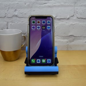 May include: A blue phone stand shaped like a chair, holding a smartphone with a blue and purple background and several app icons. The phone stand is on a wooden surface.