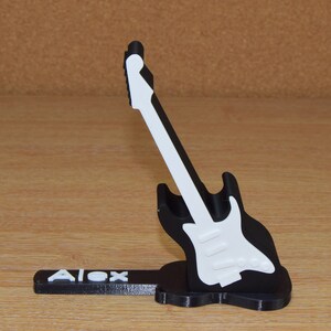 Puede incluir: Un soporte para tel&eacute;fono con forma de guitarra en blanco y negro con el nombre "Alex" impreso en la base.