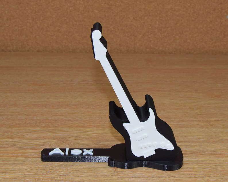 Puede incluir: Un soporte para tel&eacute;fono con forma de guitarra en blanco y negro con el nombre "Alex" impreso en la base.