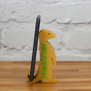 K&ouml;nnte beinhalten: Ein gelber, 3D-gedruckter Telefonst&auml;nder in Form eines Erdm&auml;nnchens. Der St&auml;nder hat das Wort "Heather" in gr&uuml;nen Buchstaben auf der Seite aufgedruckt. Ein schwarzes Smartphone befindet sich im St&auml;nder.