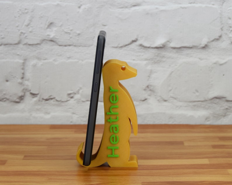 K&ouml;nnte beinhalten: Ein gelber, 3D-gedruckter Telefonst&auml;nder in Form eines Erdm&auml;nnchens. Der St&auml;nder hat das Wort "Heather" in gr&uuml;nen Buchstaben auf der Seite aufgedruckt. Ein schwarzes Smartphone befindet sich im St&auml;nder.