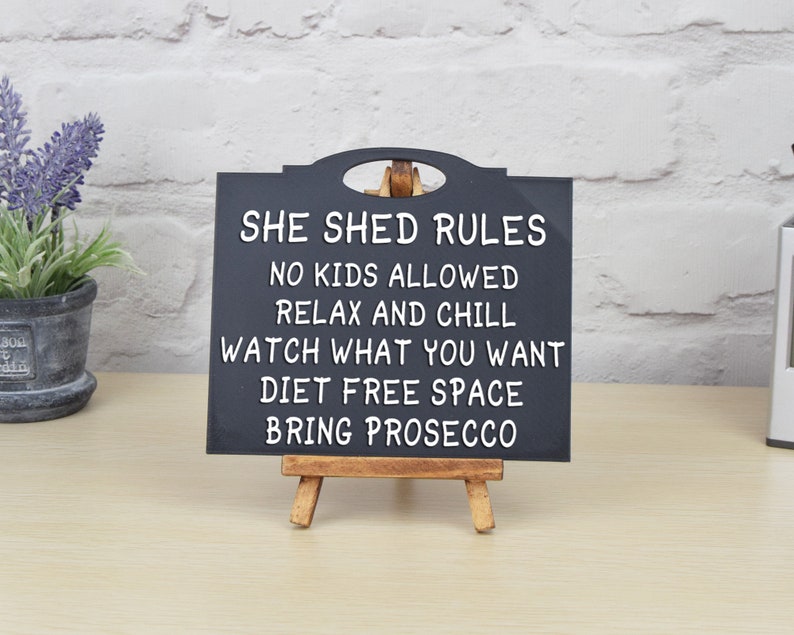 May include: Black chalkboard sign with white text that reads "She Shed Rules: No Kids Allowed, Relax and Chill, Watch What You Want, Diet Free Space, Bring Prosecco". The sign is on a small wooden easel.