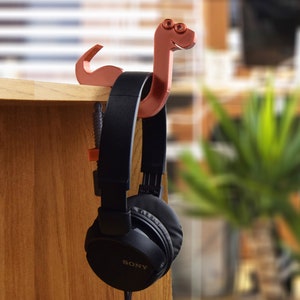 May include: A brown 3D printed snake-shaped headphone holder with a pair of black headphones hanging from it.
