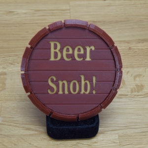 May include: A round, burgundy-colored coaster with the words "Beer Snob!" in gold lettering. The coaster is designed to resemble a wooden barrel, with horizontal lines and a circular shape.