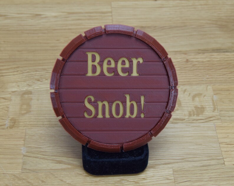 May include: A round, burgundy-colored coaster with the words "Beer Snob!" in gold lettering. The coaster is designed to resemble a wooden barrel, with horizontal lines and a circular shape.