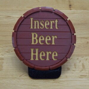 May include: A round, burgundy-colored beer coaster with the words "Insert Beer Here" in gold lettering. The coaster is designed to resemble a wooden barrel and is resting on a black base.