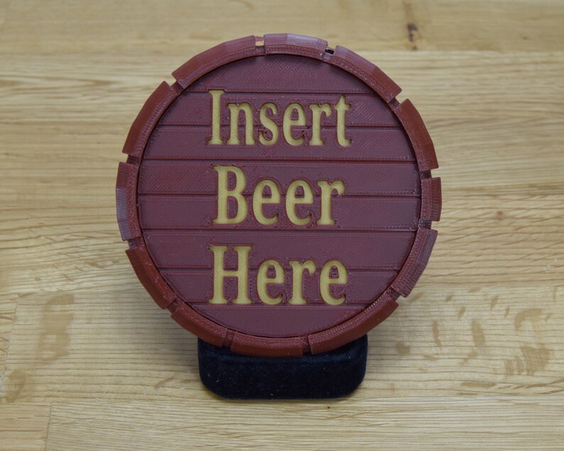 May include: A round, burgundy-colored beer coaster with the words "Insert Beer Here" in gold lettering. The coaster is designed to resemble a wooden barrel and is resting on a black base.