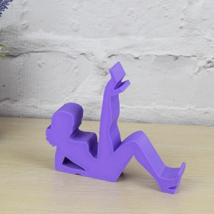 May include: Purple phone stand shaped like a person sitting with their legs crossed and holding a phone.