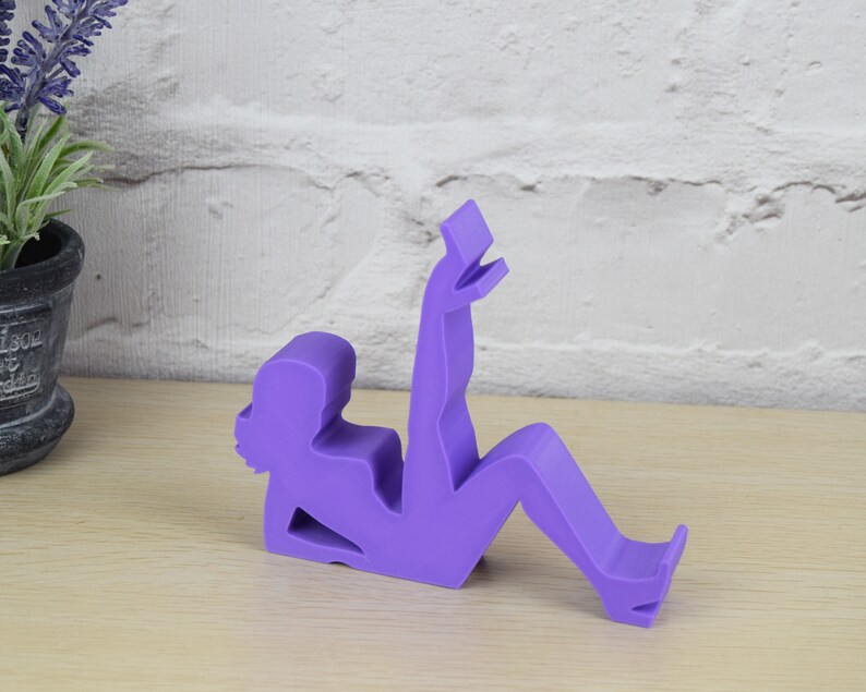 May include: Purple phone stand shaped like a person sitting with their legs crossed and holding a phone.