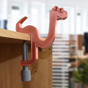 May include: A 3D printed, red-brown, snake-shaped clamp with a gray screw. The clamp is attached to a wooden surface.