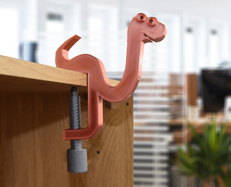 May include: A 3D printed, red-brown, snake-shaped clamp with a gray screw. The clamp is attached to a wooden surface.
