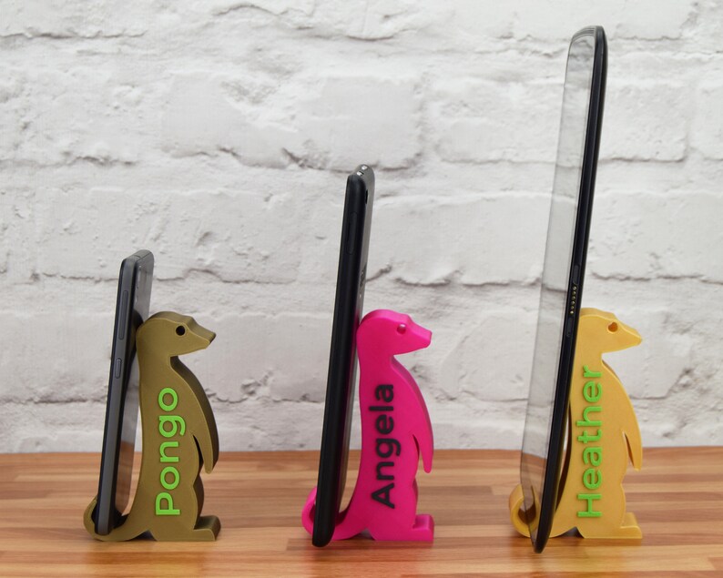 K&ouml;nnte beinhalten: Drei Handyst&auml;nder aus 3D-gedruckten Erdm&auml;nnchen, die jeweils ein Handy in unterschiedlicher Gr&ouml;&szlig;e halten. Die St&auml;nder sind gold, pink und braun. Die Namen "Pongo", "Angela" und "Heather" sind auf den St&auml;ndern aufgedruckt.