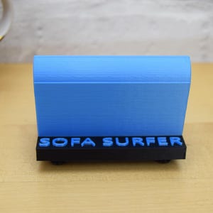 May include: A blue 3D printed object with the words "SOFA SURFER" printed on the front. The object is on a wooden surface.