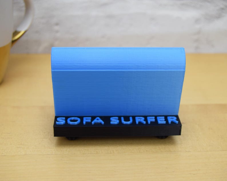 May include: A blue 3D printed object with the words "SOFA SURFER" printed on the front. The object is on a wooden surface.