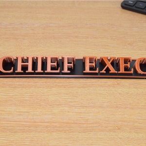May include: A black and copper desk sign with the words "CHIEF EXEC" in raised letters.