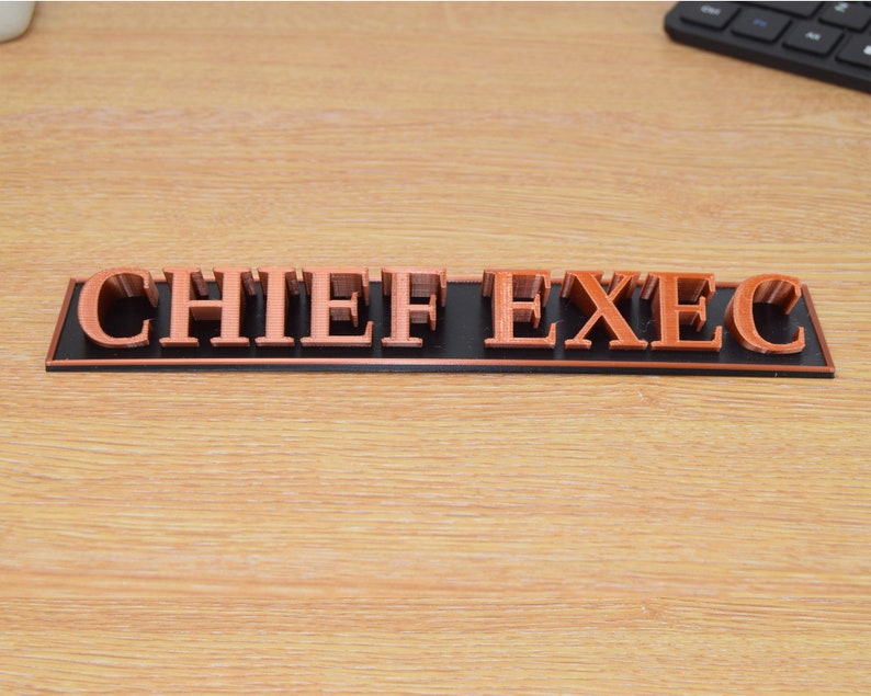 May include: A black and copper desk sign with the words "CHIEF EXEC" in raised letters.