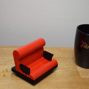 May include: A red and black 3D printed phone stand shaped like a couch. The stand has a black base and two armrests.