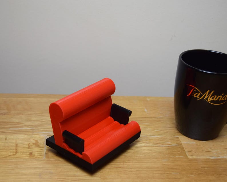 May include: A red and black 3D printed phone stand shaped like a couch. The stand has a black base and two armrests.