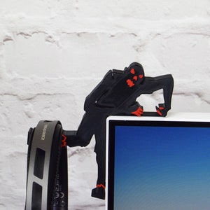May include: A black 3D printed figure of a zombie with red eyes, holding onto the top of a computer monitor. A pair of black headphones with silver accents are hanging from the monitor.