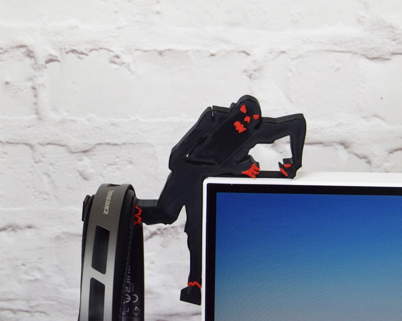 May include: A black 3D printed figure of a zombie with red eyes, holding onto the top of a computer monitor. A pair of black headphones with silver accents are hanging from the monitor.