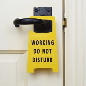 Op de afbeelding: Een geel bord met zwarte tekst dat "Working Do Not Disturb" zegt, hangt aan een witte deur.