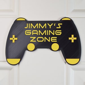 以下が含まれることがあります： 黒と黄色のゲームコントローラーのサインで、「JIMMY'S GAMING ZONE」の文字が書かれています。