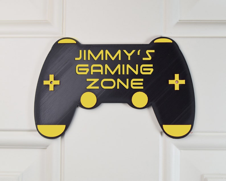 以下が含まれることがあります： 黒と黄色のゲームコントローラーのサインで、「JIMMY'S GAMING ZONE」の文字が書かれています。