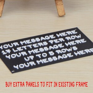 May include: Black sign with white raised letters that read "YOUR MESSAGE HERE 17 LETTERS PER ROW YOUR MESSAGE HERE UP TO 5 ROWS YOUR MESSAGE HERE". The sign is on a light wood surface. BUY EXTRA PANELS TO FIT IN EXISTING FRAME is written in red below the sign.