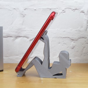 May include: A gray 3D printed phone stand shaped like a person holding a red smartphone.