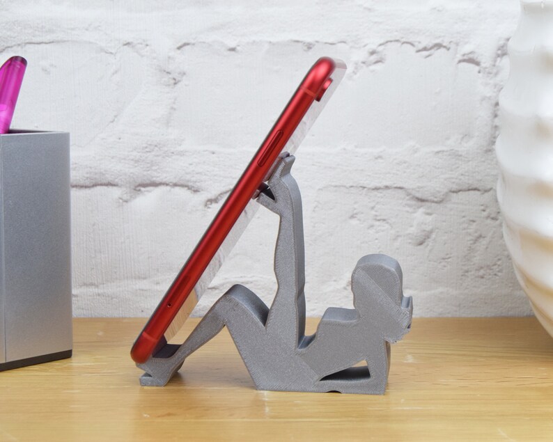 May include: A gray 3D printed phone stand shaped like a person holding a red smartphone.