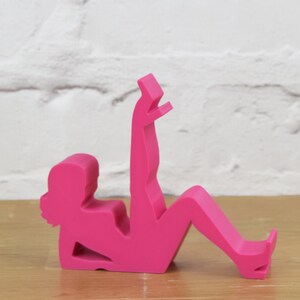 May include: A pink plastic figurine of a person in a sitting position with one leg raised. The figurine is designed to hold a phone or other device.