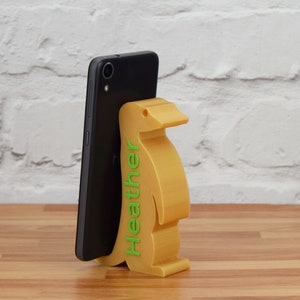 K&ouml;nnte beinhalten: Ein gelber, pinguin-f&ouml;rmiger Handyst&auml;nder mit dem Namen "Heather" auf der Seite. Ein schwarzes Smartphone befindet sich im St&auml;nder.