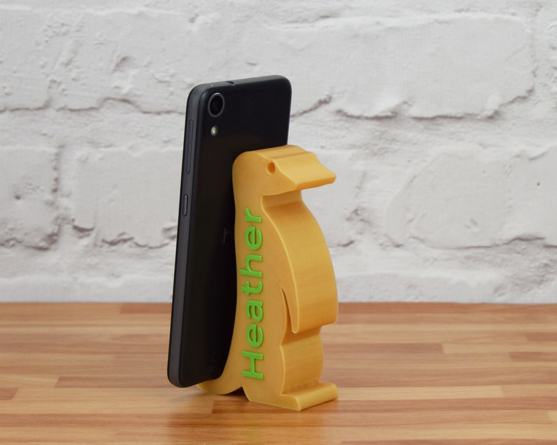 K&ouml;nnte beinhalten: Ein gelber, pinguin-f&ouml;rmiger Handyst&auml;nder mit dem Namen "Heather" auf der Seite. Ein schwarzes Smartphone befindet sich im St&auml;nder.