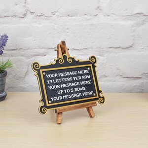 May include: A black sign with gold trim and an easel stand. The sign has the text "YOUR MESSAGE HERE 17 LETTERS PER ROW YOUR MESSAGE HERE UP TO 5 ROWS YOUR MESSAGE HERE" on it.