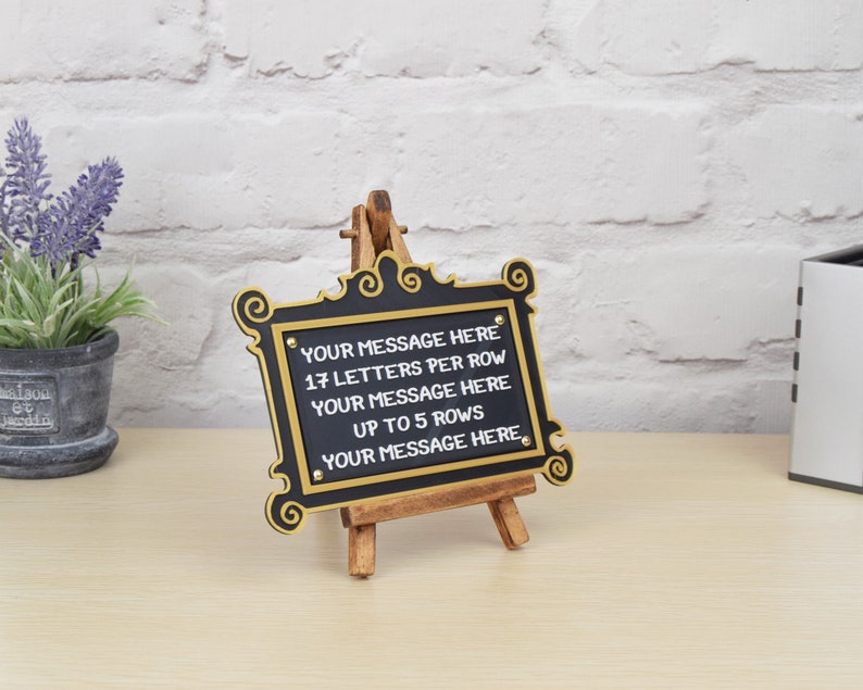 May include: A black sign with gold trim and an easel stand. The sign has the text "YOUR MESSAGE HERE 17 LETTERS PER ROW YOUR MESSAGE HERE UP TO 5 ROWS YOUR MESSAGE HERE" on it.