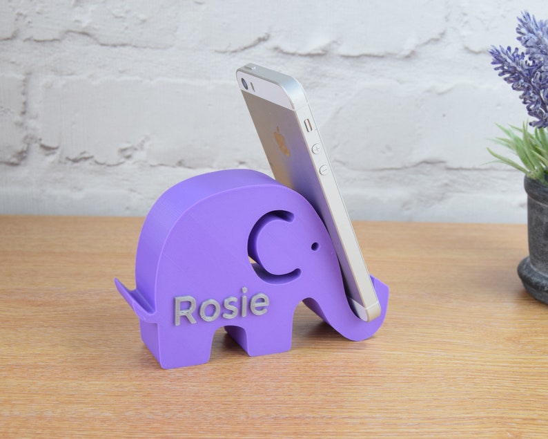 May include: Purple elephant phone stand with the name "Rosie" and the letter "C" on the side. The stand is holding a silver smartphone.